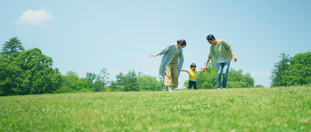 青空の下、芝生広場で親子三人が手をつなぎ、歩きながら笑顔で過ごしている様子