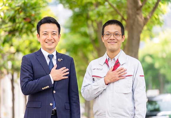 スーツ姿の男性と作業服姿の男性が、緑の木々の前で胸に手を当て、笑顔でカメラを見ている様子
