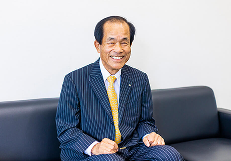 ストライプ柄のスーツ姿の男性が黒いソファに座り、笑顔で正面を向いている様子