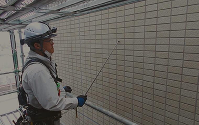 ヘルメットとハーネスを装着した作業員が、タイル張りの外壁を専用の道具で点検している様子