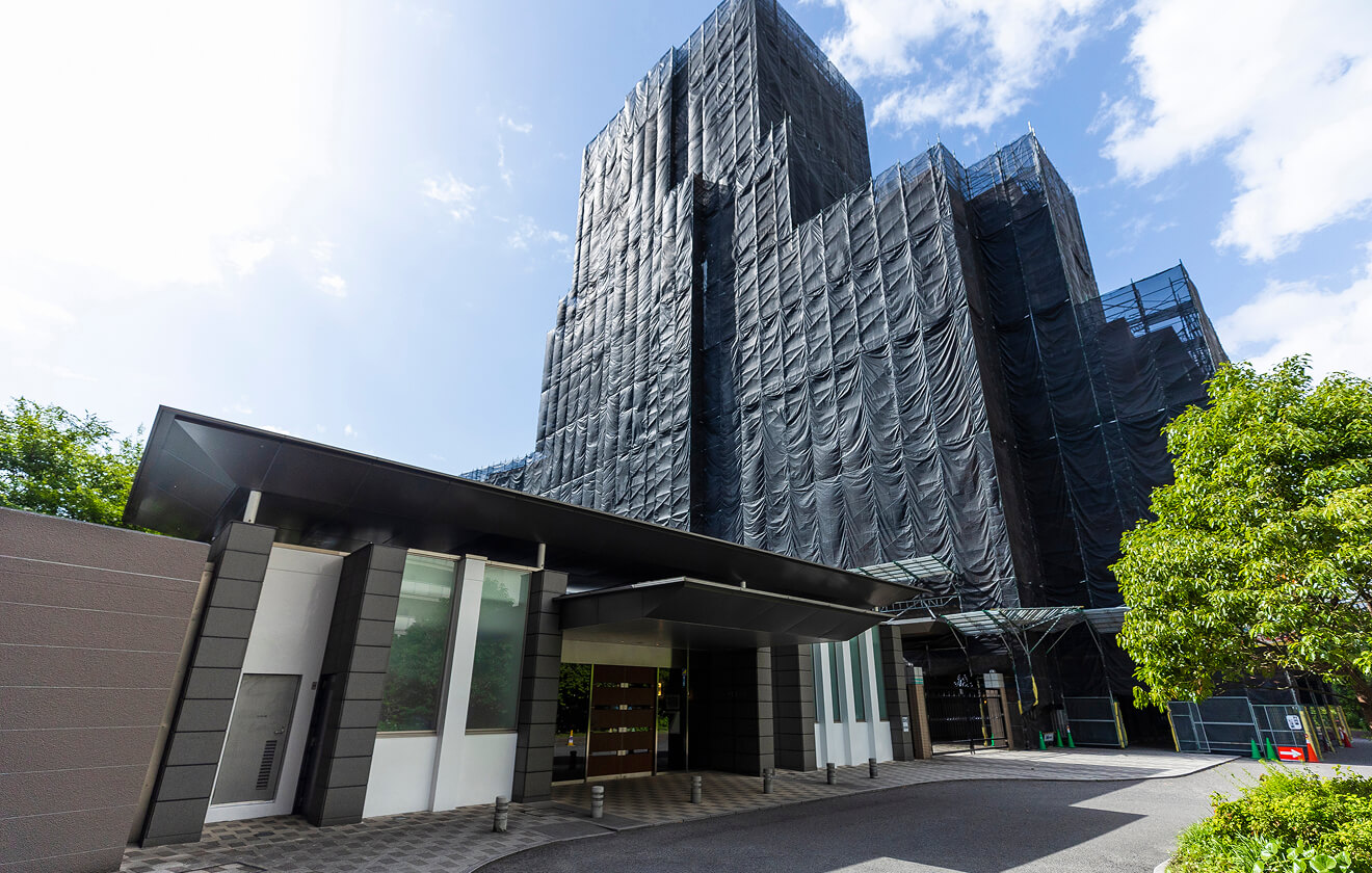 足場と養生シートで全面を覆った建物が奥に、手前にはエントランス周辺が写る修繕現場の様子