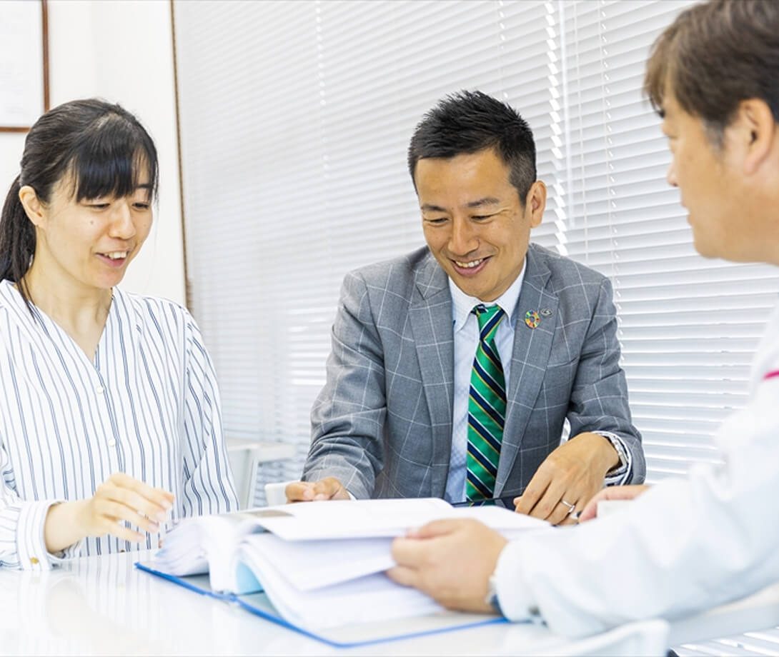 オフィスのテーブルで分厚い資料を囲み、笑顔で打ち合わせをする男女3人の社員の様子