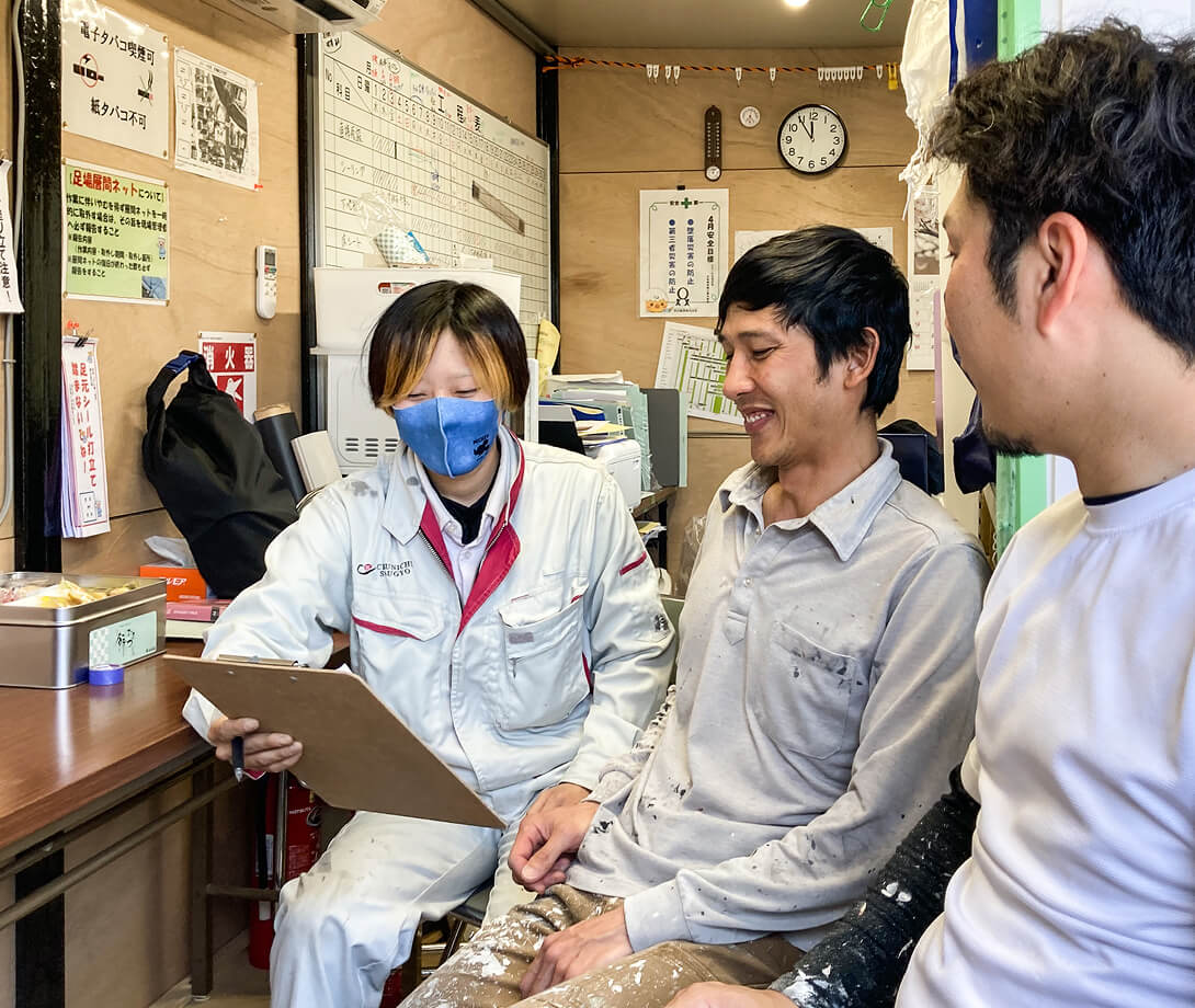 現場事務所で、クリップボードの資料を見ながら笑顔で話し合う3人の作業員の様子