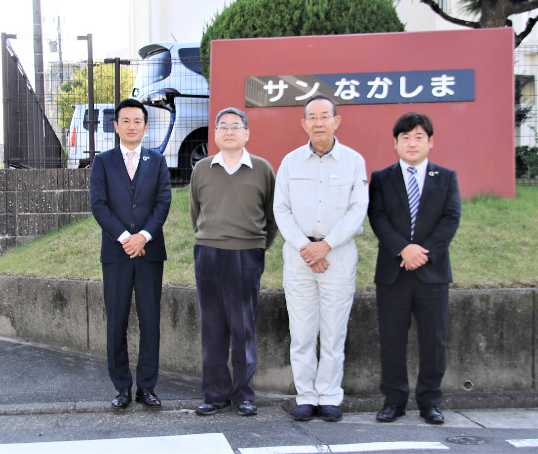 施設の赤い壁の看板の前で、スーツや作業服を着たスタッフとお客様が並んで立っている様子